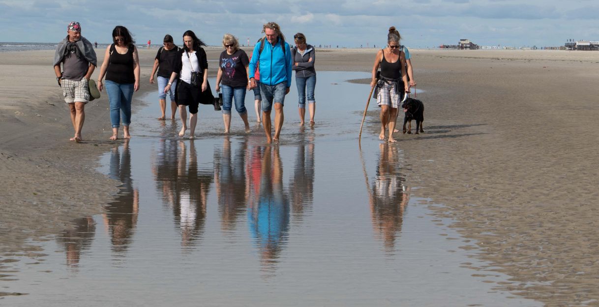 A group of people is walking barefoot along the beach. In the foreground, their reflections can be seen in the water.