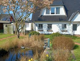 Haus mit dunklem Dach, blühendem Magnolienbaum und Gartenteich mit Schilf und Narzissen im Vordergrund.