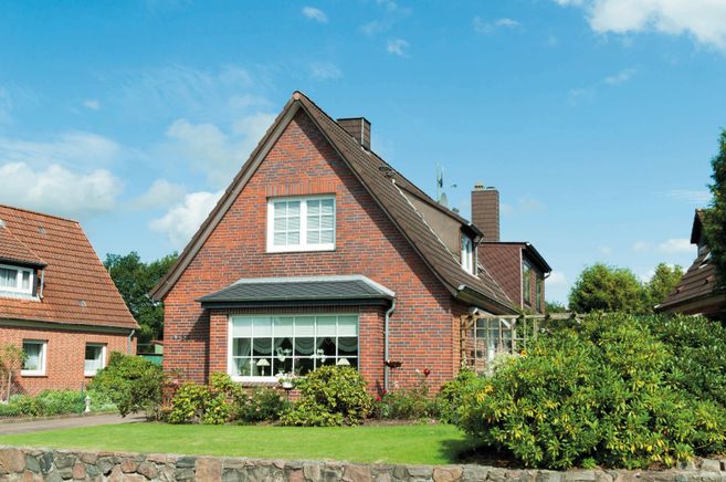 Außenansicht eines roten Backsteinhauses mit steilem Dach, Garten und blauem Himmel.