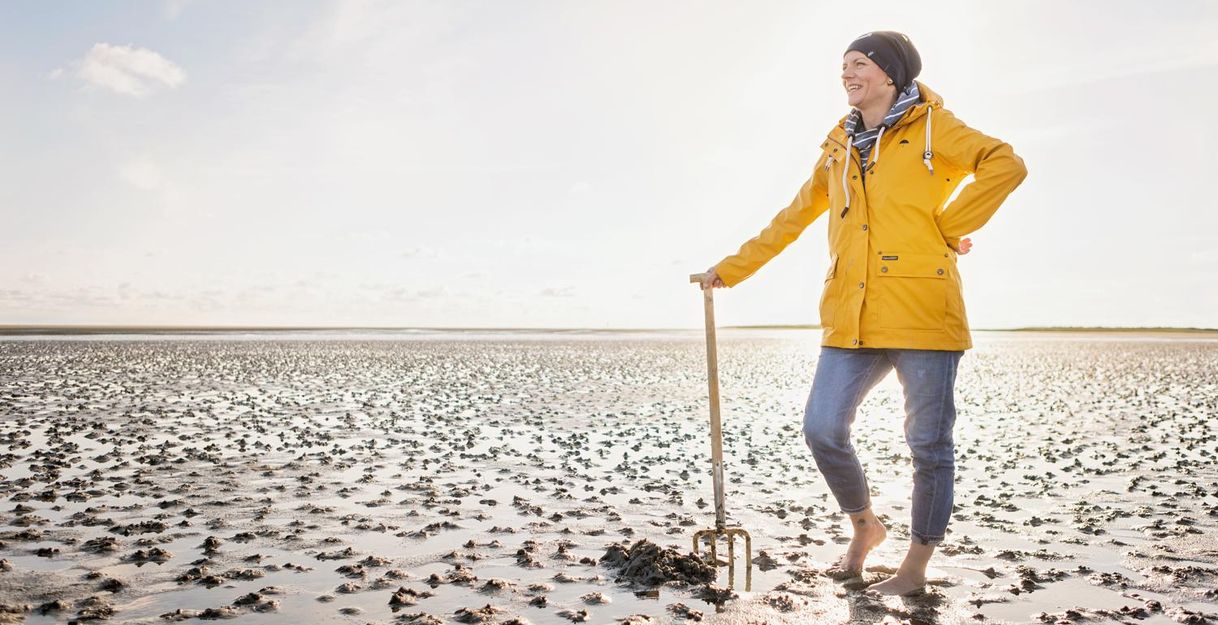 Eine Person in einem gelben Regenmantel steht barfuß am Watt. Im Hintergrund sind weite, schlickige Flächen und ein klarer Himmel zu sehen.