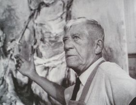 Ein älterer Mann steht vor einem großen Gemälde und gestikuliert mit der Hand.