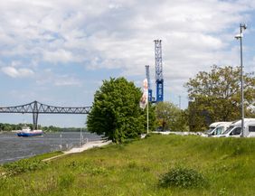 Wohnmobile stehen auf einem grünen Hügel am Nord-Ostsee-Kanal, im Hintergrund eine Brücke und ein vorbeifahrendes Schiff.