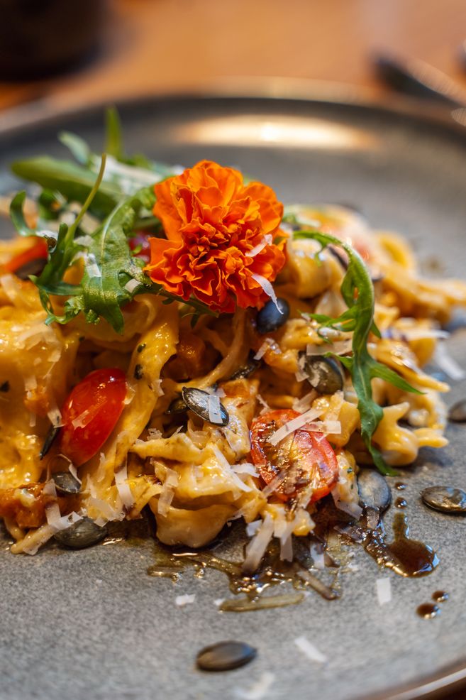 Ein Teller mit einer kulinarischen Pasta-Kreation, garniert mit einer orangefarbenen Blüte, Rucola, Kürbiskernen und Kirschtomaten.