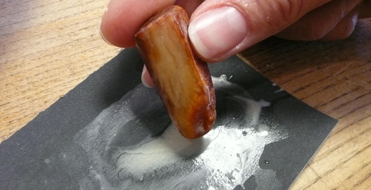 A hand is rubbing a piece of wood on a black sandpaper. There are traces of sanding dust visible.