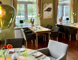 Ein gemütlich eingerichtetes Restaurant mit gedeckten Tischen, grünen Wänden und großen Fenstern. Kunstwerke und Dekorationen sind an den Wänden zu sehen.