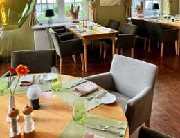 Ein gemütlich eingerichtetes Restaurant mit gedeckten Tischen, grünen Wänden und großen Fenstern. Kunstwerke und Dekorationen sind an den Wänden zu sehen.