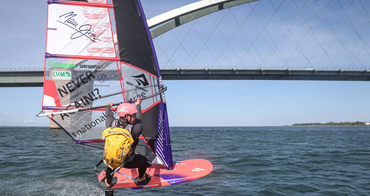 Ein Windsurfer mit gelbem Rucksack surft vor einer Brücke auf Fehmarn.