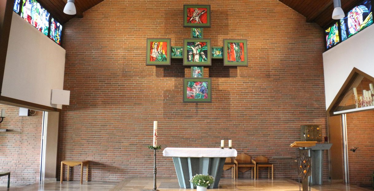 Ein Innenraum einer Kirche mit einem schlichten Altar und bunten Fenstern. Die Wände sind aus Backstein und es gibt eine Kreuzsymbolik an der Rückwand.