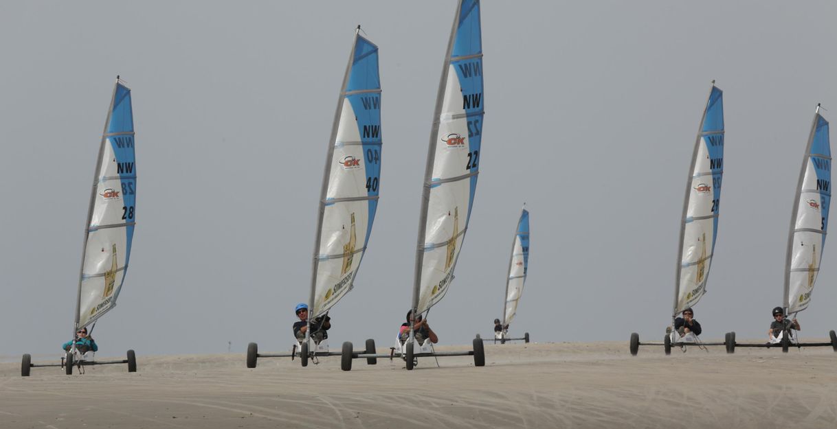 Six land sailers drive over a sandy hill. The colorful sails stand out against the gray sky.