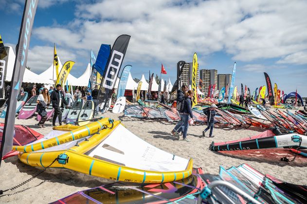 Menschen auf einem Strand mit vielen Surfsegeln und bunten Fahnen beim Surf Festival 2023.