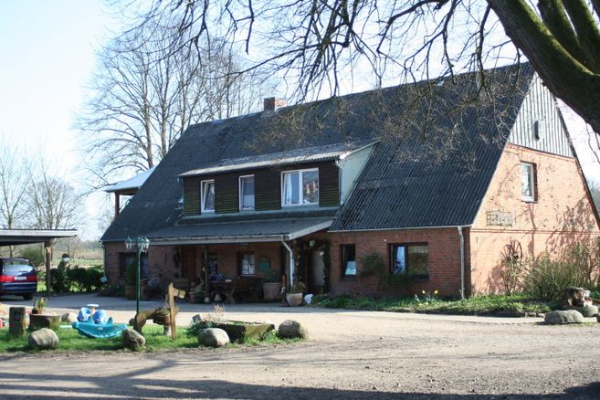 Landhaus mit großem, steilem Dach, roter Backsteinfassade und Vorgarten; links steht ein Auto, im Vordergrund Spielzeug und Steine.