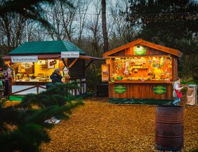 A Christmas market with wooden stalls outdoors. The booths offer various goods and foods, surrounded by trees and festive lighting.