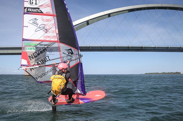 Ein Windsurfer mit gelbem Rucksack surft vor einer Brücke auf Fehmarn.