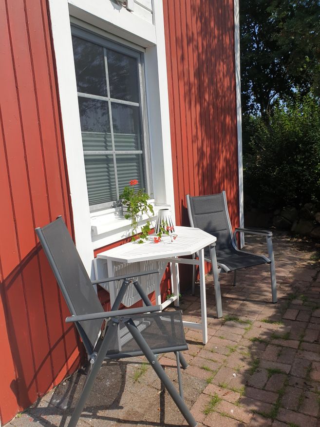 Terrasse mit zwei grauen Stühlen, weißem Tisch und roter Holzfassade mit Fenster im Sonnenlicht.