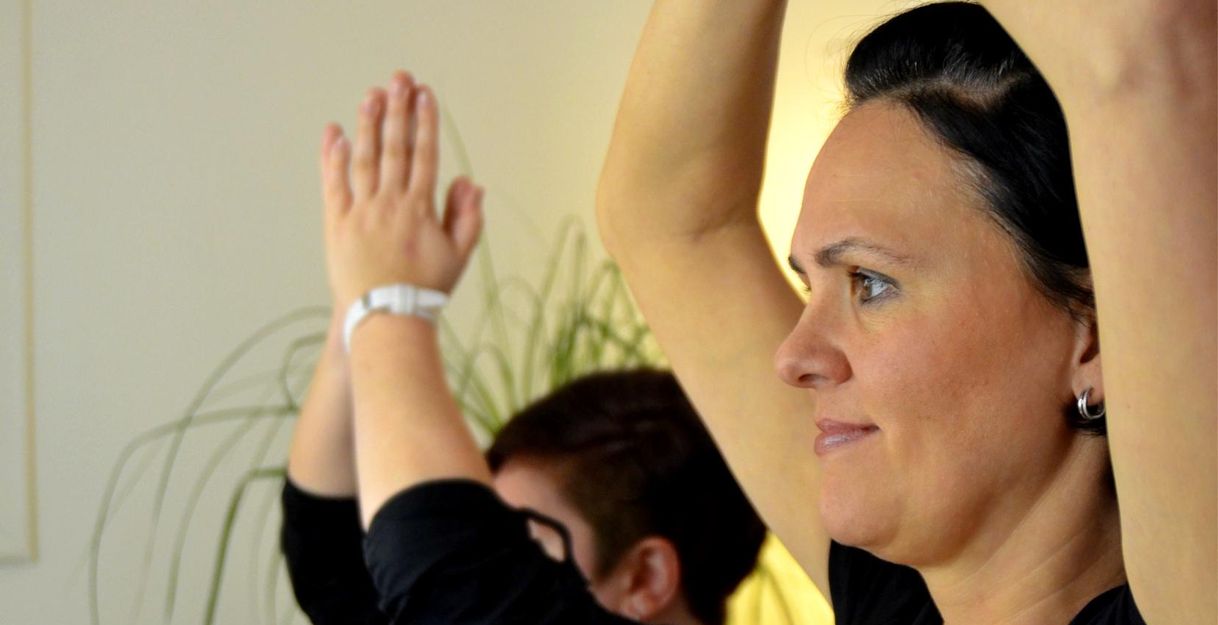 Zwei Frauen in schwarzen T-Shirts üben eine Yoga-Position mit erhobenen Händen. Der Raum ist hell und bietet eine entspannte Atmosphäre.