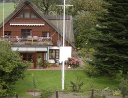 Ein rotes Reetdachhaus, umgeben von Bäumen und einem Garten. Im Vordergrund grasen Schafe auf einer Wiese.