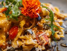 Ein Teller mit einer kulinarischen Pasta-Kreation, garniert mit einer orangefarbenen Blüte, Rucola, Kürbiskernen und Kirschtomaten.