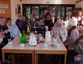 A group of elderly people is sitting at tables in a bright room. They are happily chatting over coffee and cake.