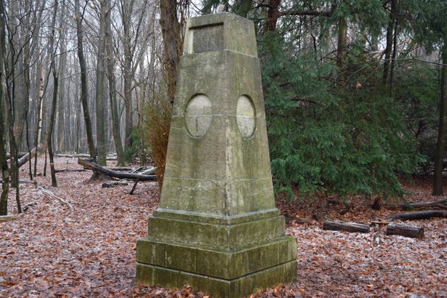 Altes steinernes Denkmal im herbstlich-winterlichen Wald bei Schafflund. Der Obelisk steht auf einem Sockel, seine Oberfläche ist verwittert. Auf der Vorderseite befinden sich zwei ovale Vertiefungen und eine verblasste Inschrift. Rundherum liegen gefallene Blätter und vereinzelte Äste, der Wald wirkt ruhig und naturbelassen.