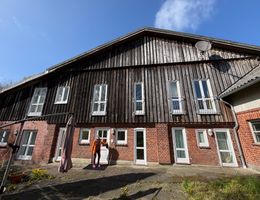 Ein zweistöckiges Gebäude mit einer Fassade aus Holz und Backstein. Eine Person steht vor der Eingangstür. Der Himmel ist blau und sonnig.