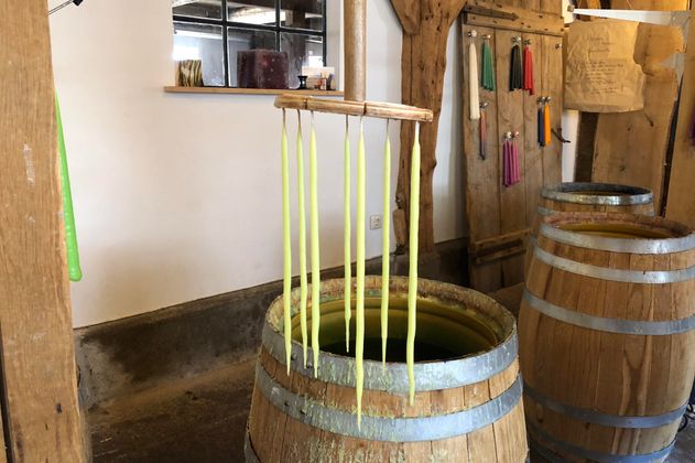 A traditional candle making workshop with several wax strands hanging from a wooden frame. In the background, wooden barrels and colorful wax strings are visible.
