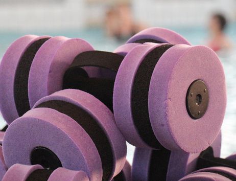 Floating dumbbells in purple color are lying on the edge of the pool. In the background, a swimming pool can be seen.