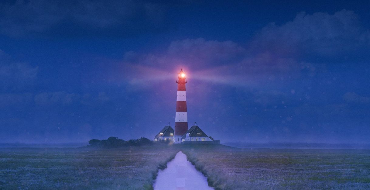 A lighthouse with red and white stripes shines in the night. Surrounded by a foggy landscape, a narrow path leads to the lighthouse.