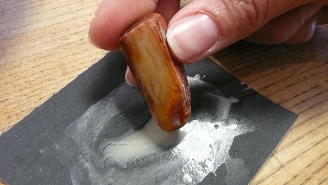 Ein Hand hält ein Stück Holz, das auf einem schwarzen Schleifpapier reibt. Es sind Spuren von Schleifstaub sichtbar.