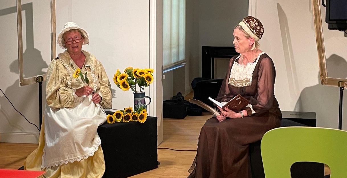 Zwei Frauen in historischen Kostümen auf einer Theaterbühne mit Sonnenblumen.