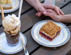 Ein Kuchen und eine Eisschokolade liegen auf einem Tisch. Zwei Hände liegen daneben.