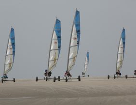Six land sailers drive over a sandy hill. The colorful sails stand out against the gray sky.
