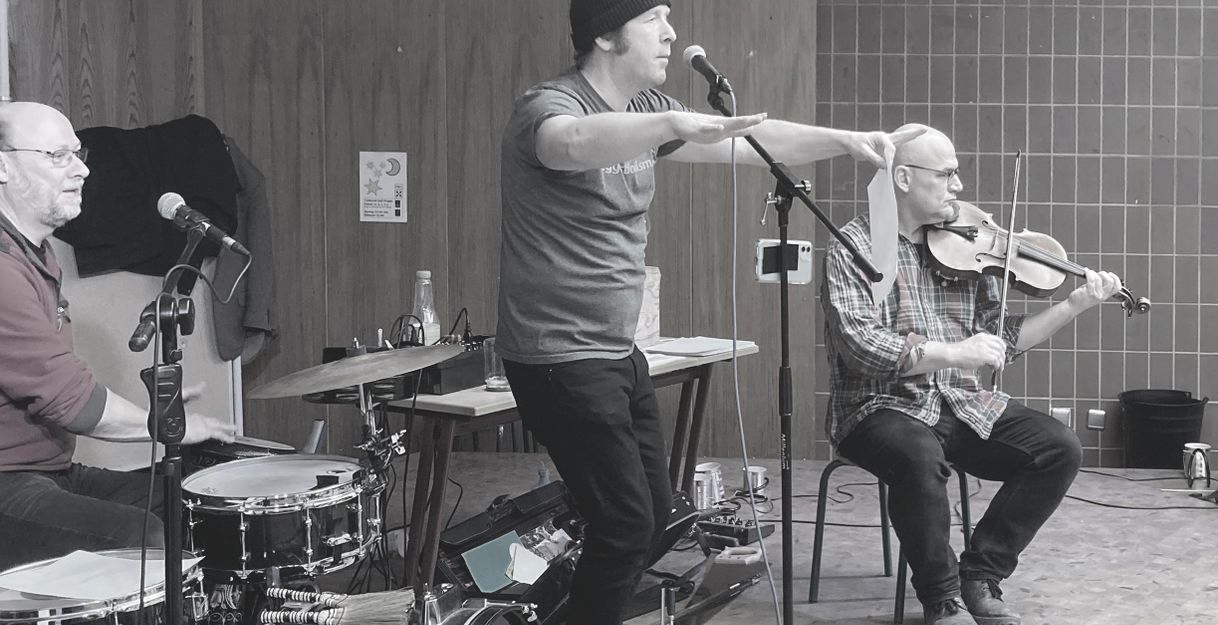 Drei Musiker bei einer Probe: ein Schlagzeuger, ein Sänger und ein Geiger.