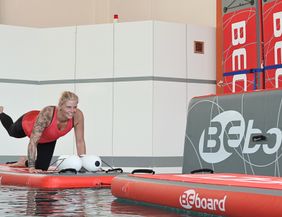 Eine Frau macht in Sportkleidung Übungen auf einem Brett in einem Schwimmbecken. 
