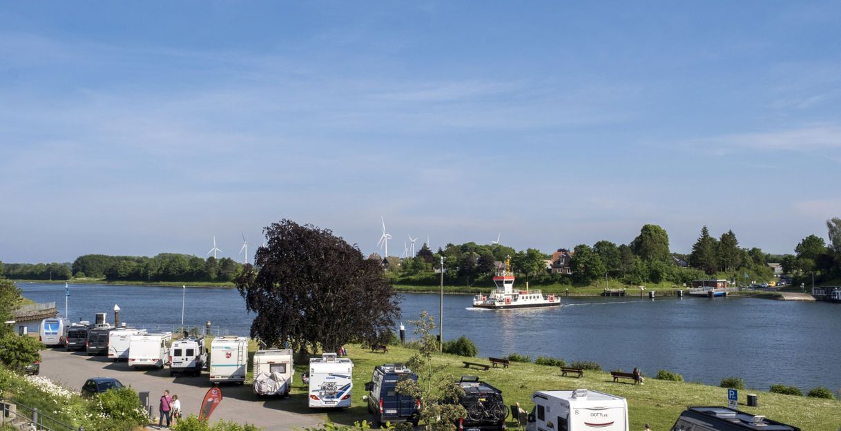 Wohnmobilstellplatz am Wasser mit Fähre und Windrädern im Hintergrund.