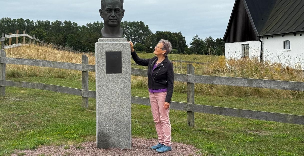 Eine Frau steht neben einer Büste auf einem Sockel im Freien, umgeben von einem Holzzaun und einem Gebäude im Hintergrund.