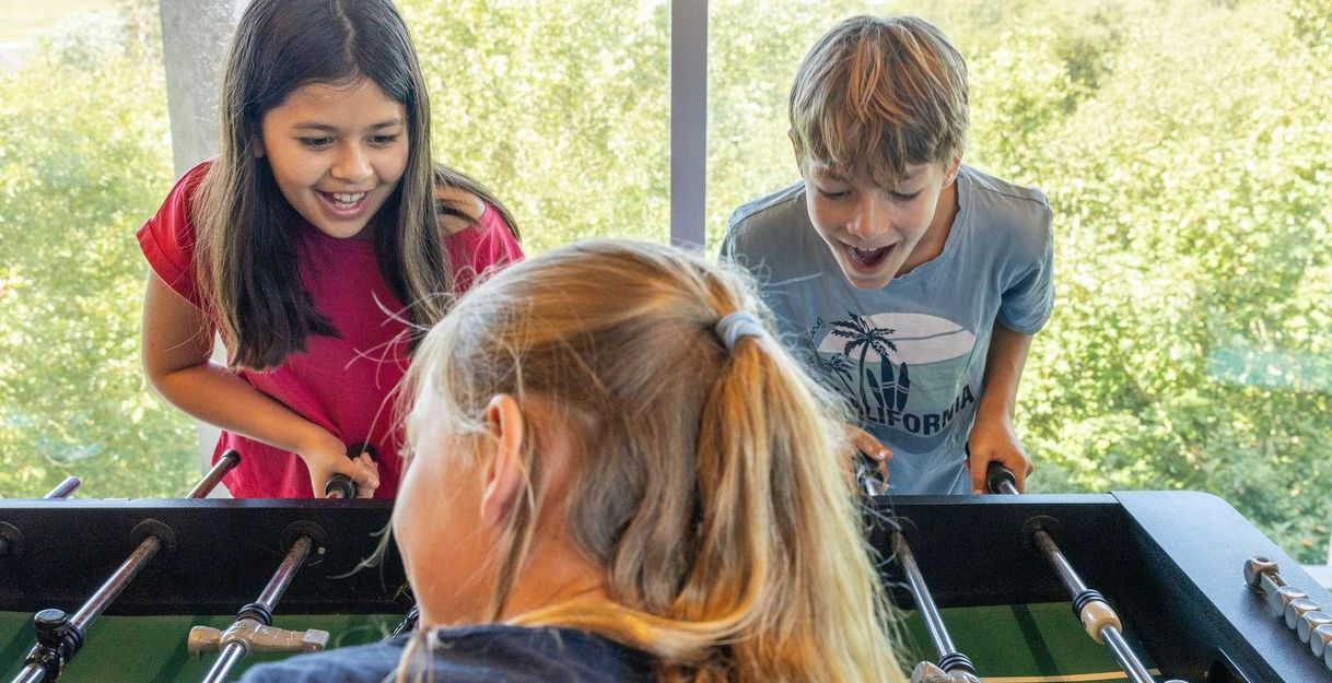 Drei Kinder spielen begeistert an einem Tischkicker. Sie lachen und haben Freude an ihrem Spiel.