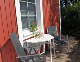Terrasse mit zwei grauen Stühlen, weißem Tisch und roter Holzfassade mit Fenster im Sonnenlicht.