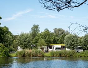 Wohnmobilstellplatz am Flussufer mit Bäumen und Wohnmobilen im Hintergrund.
