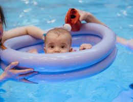 Ein Baby in einem lilafarbenen Schwimmring im Pool, gehalten von einer erwachsenen Person.