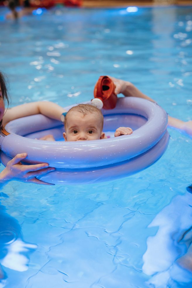 Ein Baby in einem lilafarbenen Schwimmring im Pool, gehalten von einer erwachsenen Person.