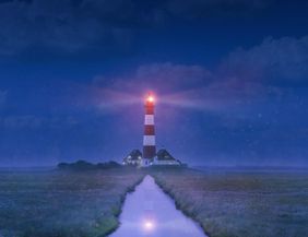 A lighthouse with red and white stripes shines in the night. Surrounded by a foggy landscape, a narrow path leads to the lighthouse.
