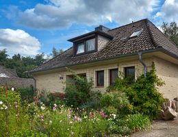Haus mit Ziegeldach und blühendem Vorgarten unter blauem Himmel mit Wolken.