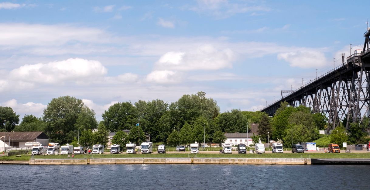 Wohnmobile parken am Ufer eines Flusses mit einer großen Brücke im Hintergrund.