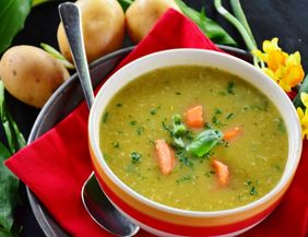 A bowl of creamy soup, garnished with fresh herbs and vegetables. Next to it are potatoes and fresh flowers on a red cloth.