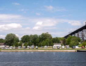 Wohnmobile parken am Ufer eines Flusses mit einer großen Brücke im Hintergrund.