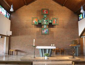 Ein Innenraum einer Kirche mit einem schlichten Altar und bunten Fenstern. Die Wände sind aus Backstein und es gibt eine Kreuzsymbolik an der Rückwand.