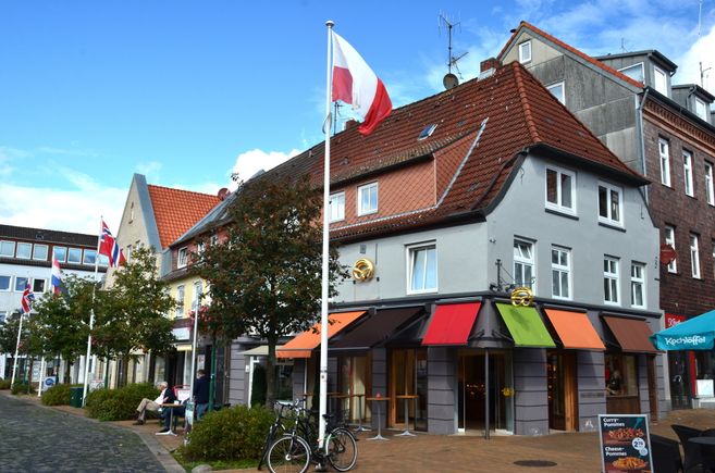 Außenansicht eines Hauses mit rotem Ziegeldach, bunten Markisen und mehreren Flaggen an einem Platz.