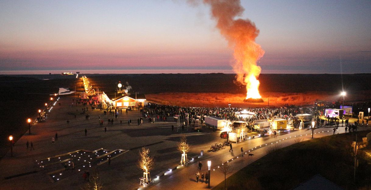 Ein großes Feuer mit rauchenden Flammen brennt am Abend. Viele Menschen versammeln sich auf dem Weg in der Nähe des Feuers.