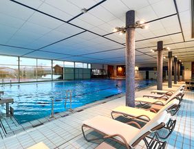 Innenansicht eines Hallenbads mit Schwimmbecken und Liegestühlen.