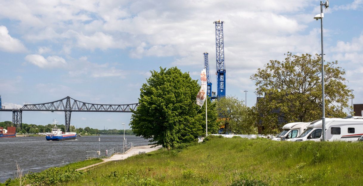 Wohnmobile stehen auf einem grünen Hügel am Nord-Ostsee-Kanal, im Hintergrund eine Brücke und ein vorbeifahrendes Schiff.